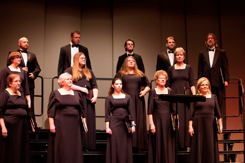 A Chior stading on risers singing, they are both men and women wearing tuxedos and dresses